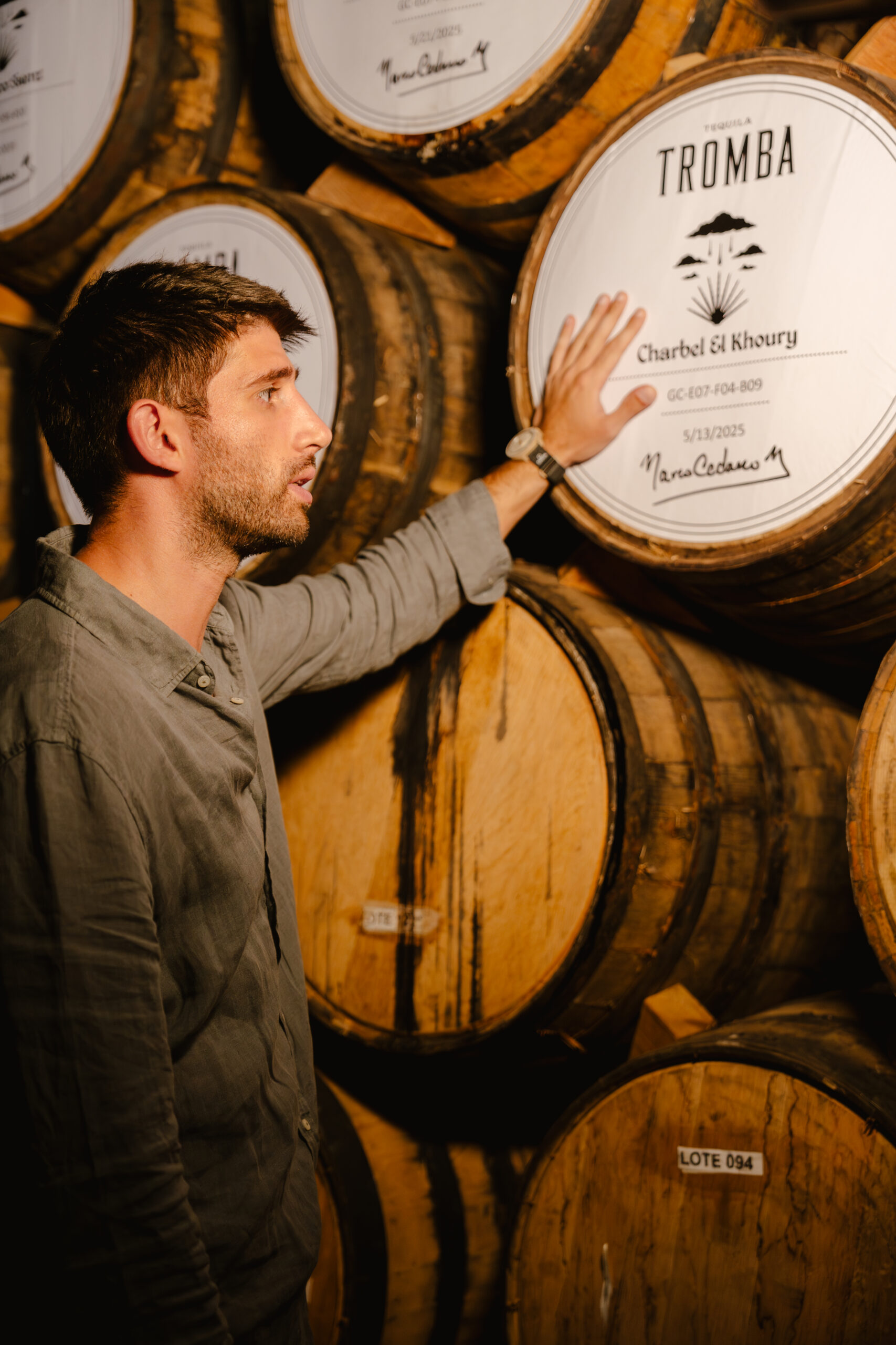 Marc Mouannes examines a labelled tequila barrel while standing among stacked oak casks in a warmly lit maturation warehouse