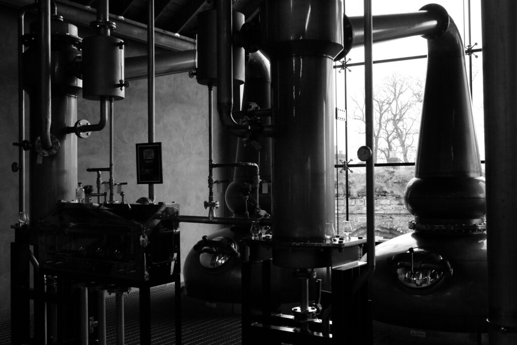Black and white photo of copper pot stills and piping inside a whisky distillery, with tasting glasses on a ledge and a large window overlooking bare trees.