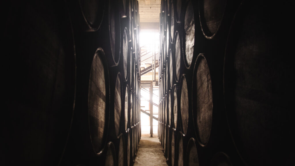 Rows of large wooden barrels line a narrow aisle inside a tequila distillery, with natural light streaming in from the far end of the tequila barrel warehouse.