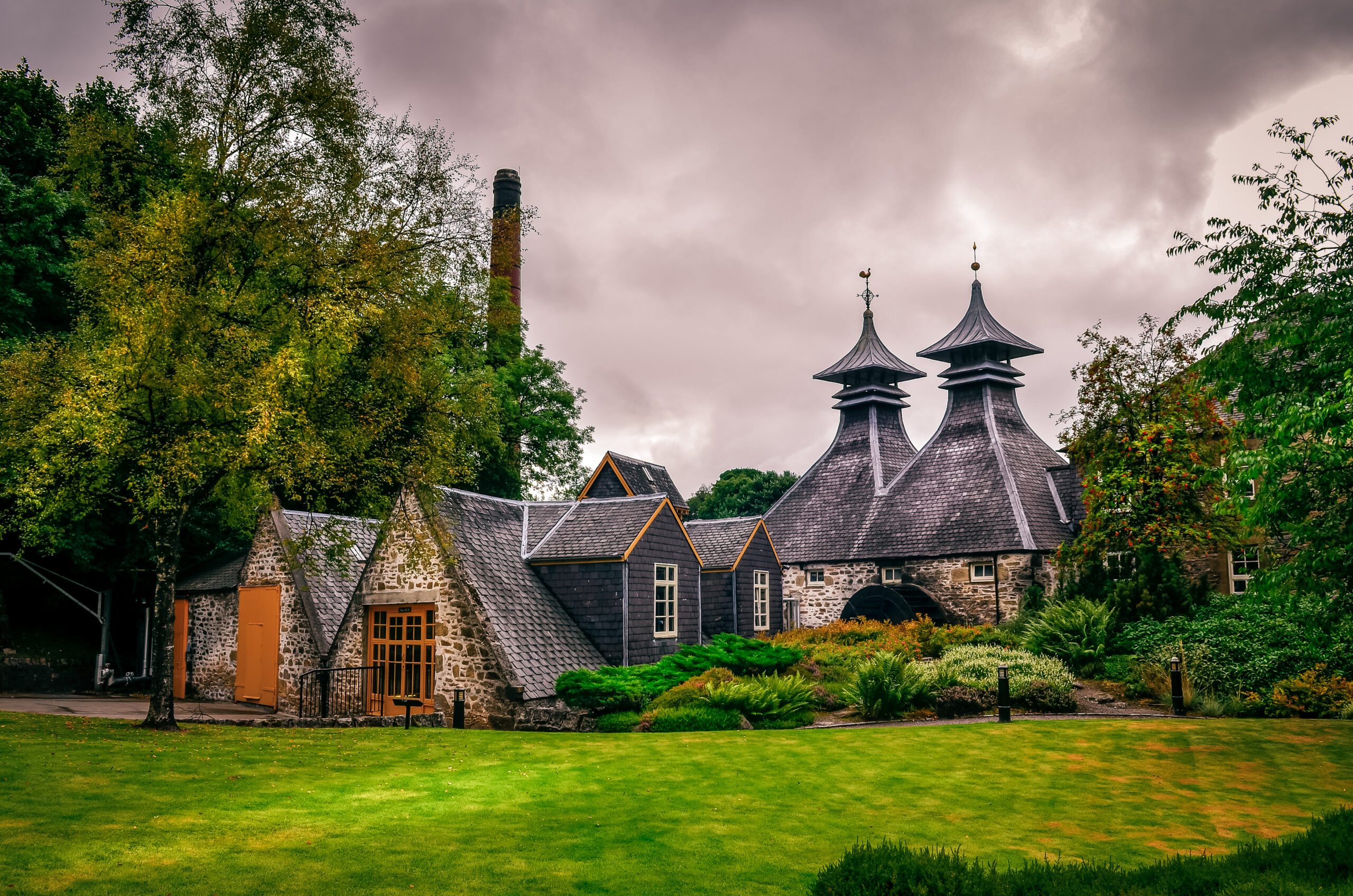 Traditional stone distillery buildings with distinctive pagoda-style roofs, surrounded by gardens and trees beneath a dramatic overcast sky.