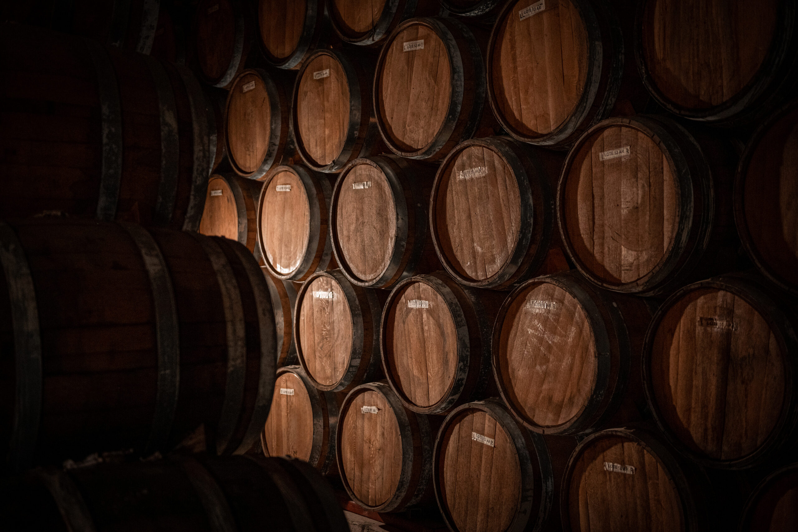 Stacked oak barrel ends in a dimly lit tequila aging cellar, each labeled "Lote 53 AR."