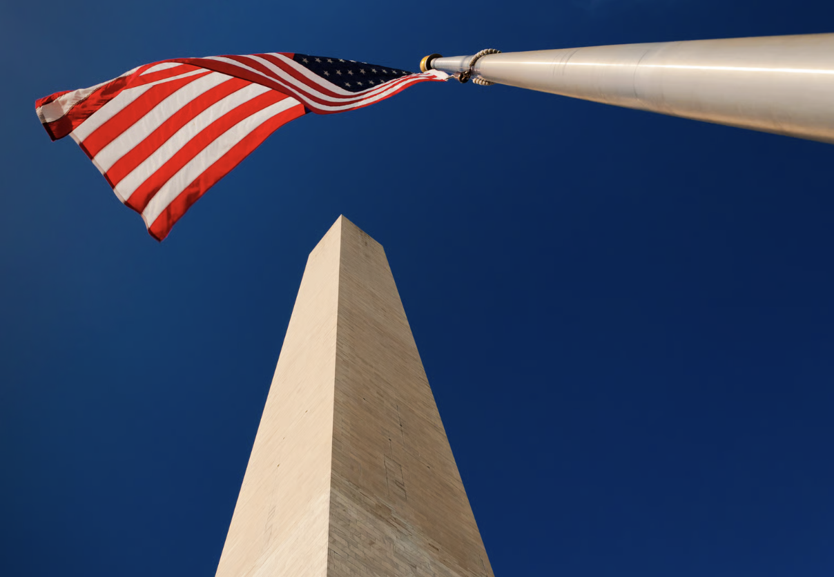 Low-angle view of an American flag flying beside the top of the Washington Monument against a deep blue sky.