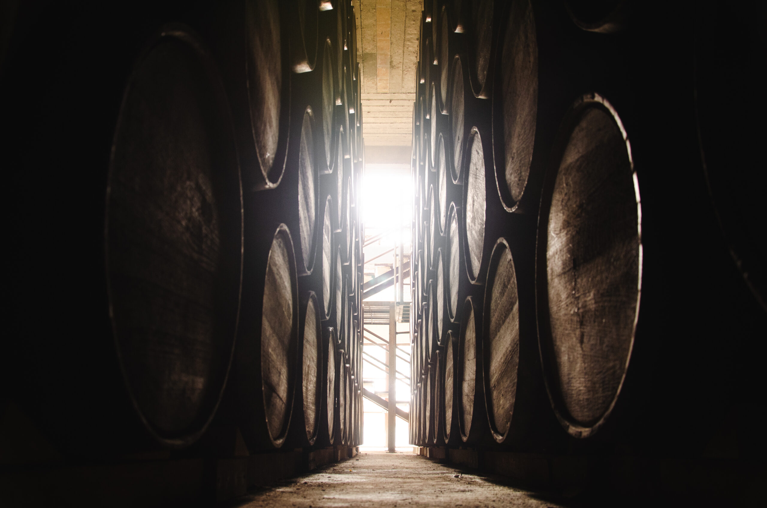 A narrow corridor formed by stacked whisky barrels, leading toward a bright light at the far end.