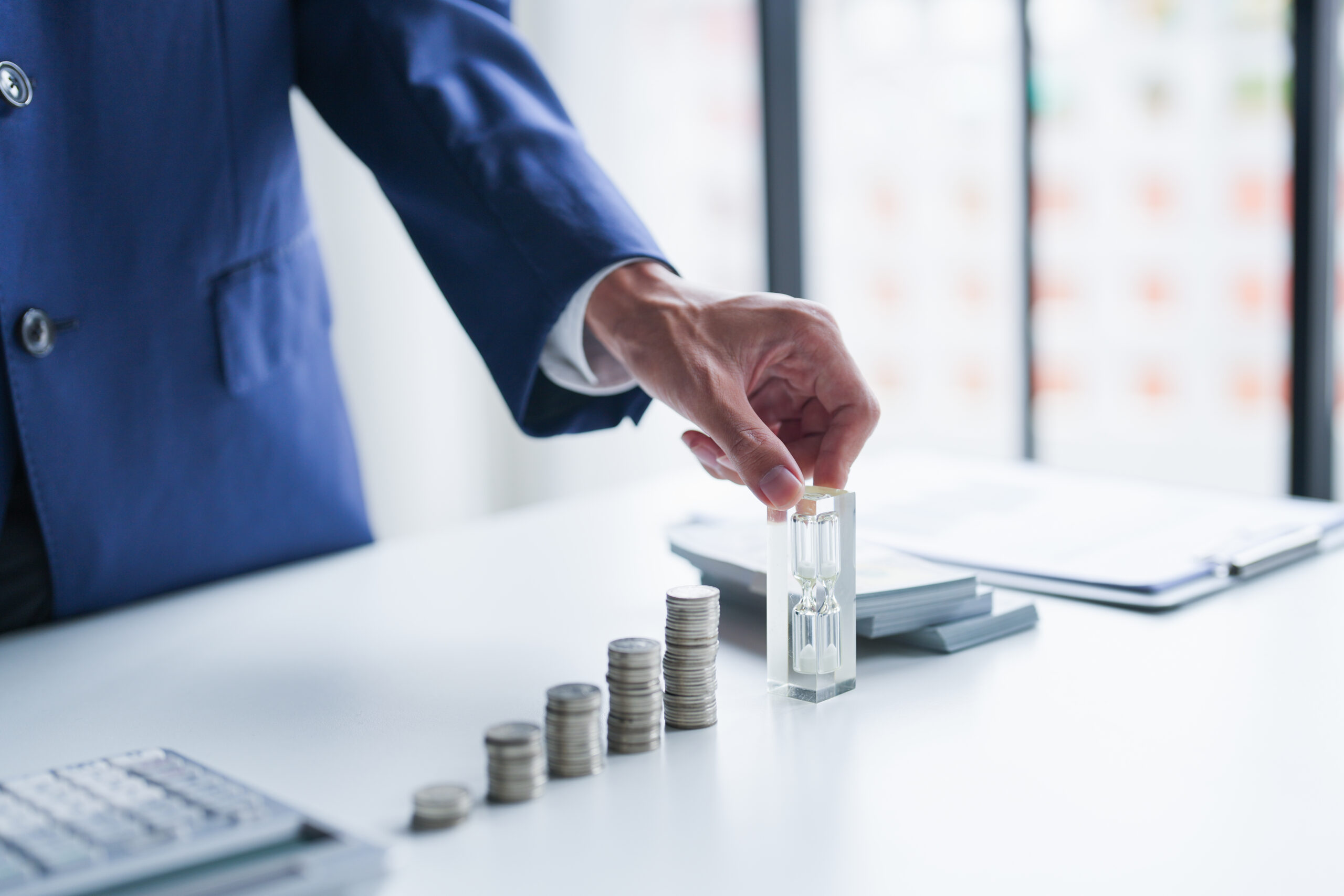 A businessman in a suit reaches for an hourglass beside ascending stacks of coins on a desk.