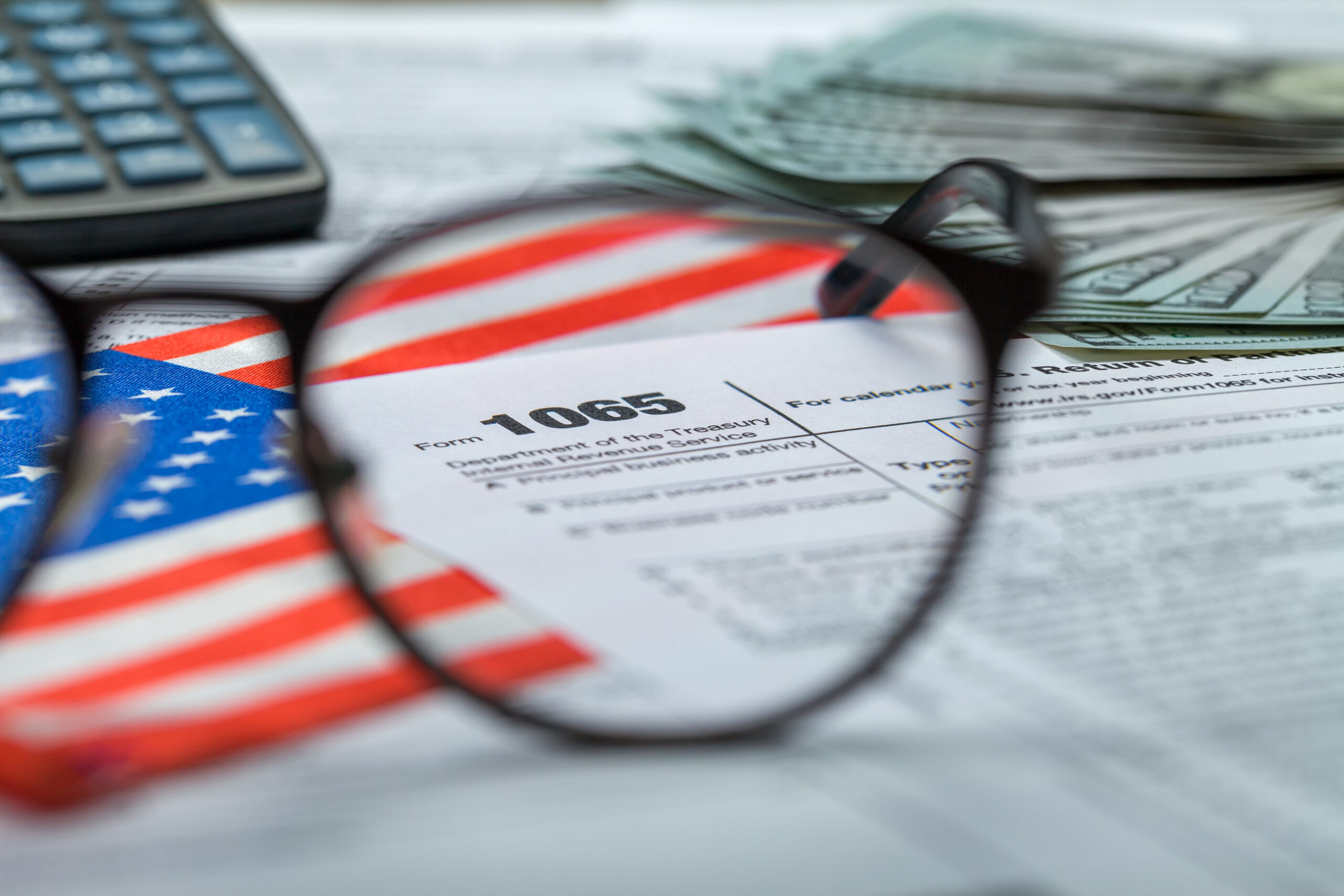 A close-up, focused shot through a pair of black-rimmed eyeglasses magnifying an IRS Form 1065, surrounded by U.S. dollar bills, an American flag, and a financial calculator.