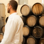 Samuel Gordon, CEO and Co-Founder of GORDON in a white shirt walks past numbered oak barrels stacked beside a 5,500-litre tank.