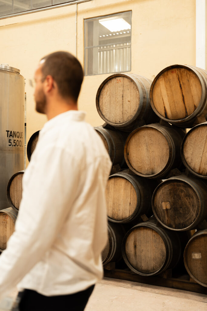 Samuel Gordon, CEO and Co-Founder of GORDON in a white shirt walks past numbered oak barrels stacked beside a 5,500-litre tank.