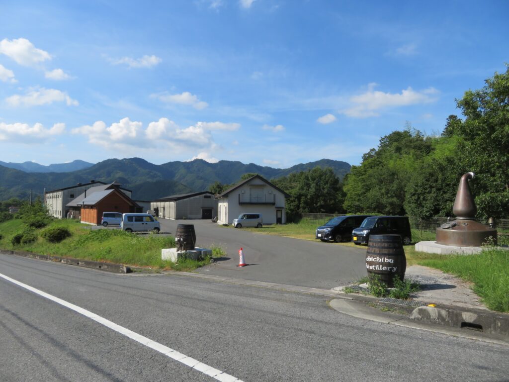 Entrance to Chichibu Distillery, with a barrel sign, a copper still monument, and distillery buildings set against forested mountains.